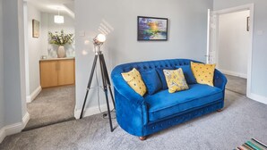 Ein blaues Samtsofa mit gelb gemusterten Kissen, eine Stehlampe und ein gerahmtes Bild an der Wand.