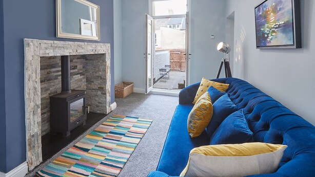 Ein Wohnzimmer mit Kamin, einem blauen Sofa mit gelben Kissen, einem farbenfrohen Teppich und einem Gemälde an der Wand.