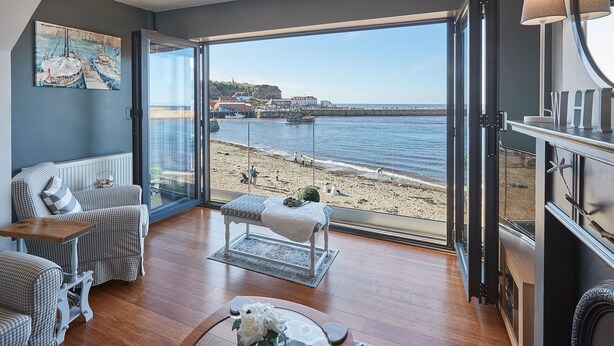 A room with a large glass door leading to a beach, wooden flooring, a checkered sofa, a small table, and a painting on the wall. - Host Stay No 3 Sandside (Whitby)