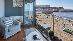A balcony with a checkered sofa, a glass door, and a view of a beach and buildings. - Host Stay No 3 Sandside (Whitby)