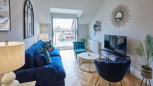Un salon moderne avec un canapé bleu, un fauteuil turquoise, une table basse ronde, un téléviseur et une plante en pot.