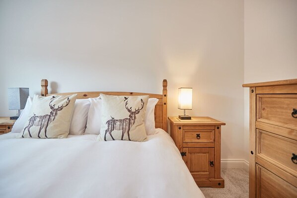 A neatly made bed with white bedding and two decorative pillows featuring deer prints, a wooden nightstand with a lamp, and a wooden dresser with multiple drawers.