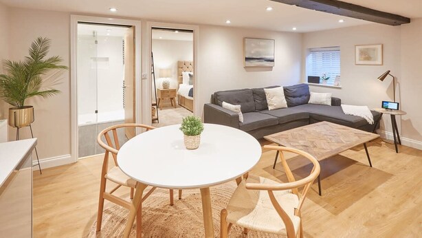 A modern living room with a grey sectional sofa, a round white table, and wooden chairs. There is a large window with blinds, a potted plant, and a painting on the wall. - Host Stay Staffordshire Hideaway (Whitby)