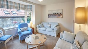A living room with a blue armchair, a white sofa, a round wooden coffee table, and a painting of a harbor scene. - Host Stay Tenby Cottage (Whitby)
