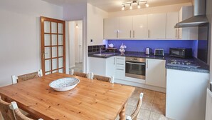 A modern kitchen with white cabinets, a wooden dining table, and a blue accent wall. - Host Stay Tenby Cottage (Whitby)
