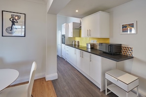 A compact kitchen with white cabinets, a microwave, and a toaster. There is a small white table and a chair in the corner.