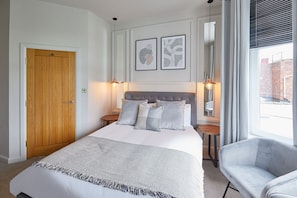 A bedroom with a bed, two bedside tables, a chair, a wooden door, and two framed artworks on the wall.