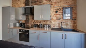 Une cuisine compacte avec des meubles gris, un four noir, un évier en acier inoxydable et un mur en briques en arrière-plan.