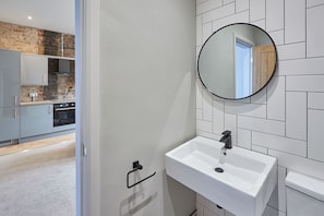 A modern bathroom with a white sink, a round mirror, and a view of a kitchen with grey cabinets and a brick wall.