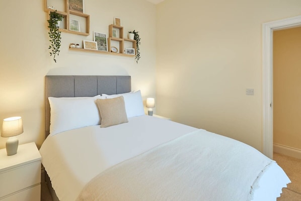 A neatly made bed with a grey headboard, white bedding, and a single decorative pillow. There are two bedside lamps on white nightstands.