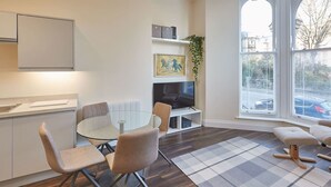 Un salon moderne avec une table à manger en verre, des chaises rembourrées, un téléviseur à écran plat et une grande fenêtre équipée de stores.