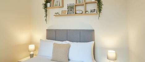 A neatly made bed with white linens, a grey upholstered headboard, and two bedside lamps. Above the bed, there are wooden shelves with framed pictures and a small clock.