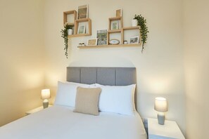 A neatly made bed with white linens, a grey upholstered headboard, and two bedside lamps. Above the bed, there are wooden shelves with framed pictures and a small clock.