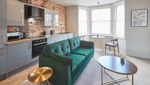 Una cocina compacta con pared de ladrillo, sofá verde y un pequeño comedor con mesa y sillas redondas.