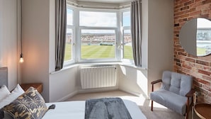 Habitación de hotel con un ventanal grande, una cama, una silla, un radiador y un espejo en la pared de ladrillo.