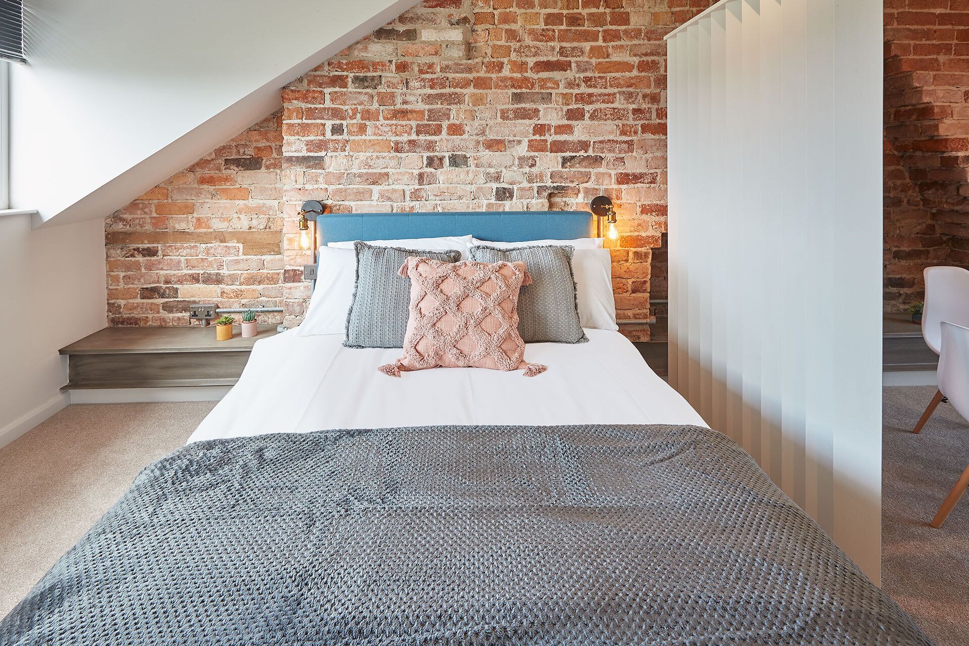 A bedroom with a brick wall, a sloped ceiling, and a bed with multiple pillows and a blanket.