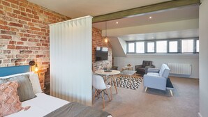 Une chambre d’hôtel moderne avec un mur en briques, un lit doté d’une tête de lit bleue, un rideau blanc, une petite table ronde avec des chaises, un canapé et une grande fenêtre avec des stores.