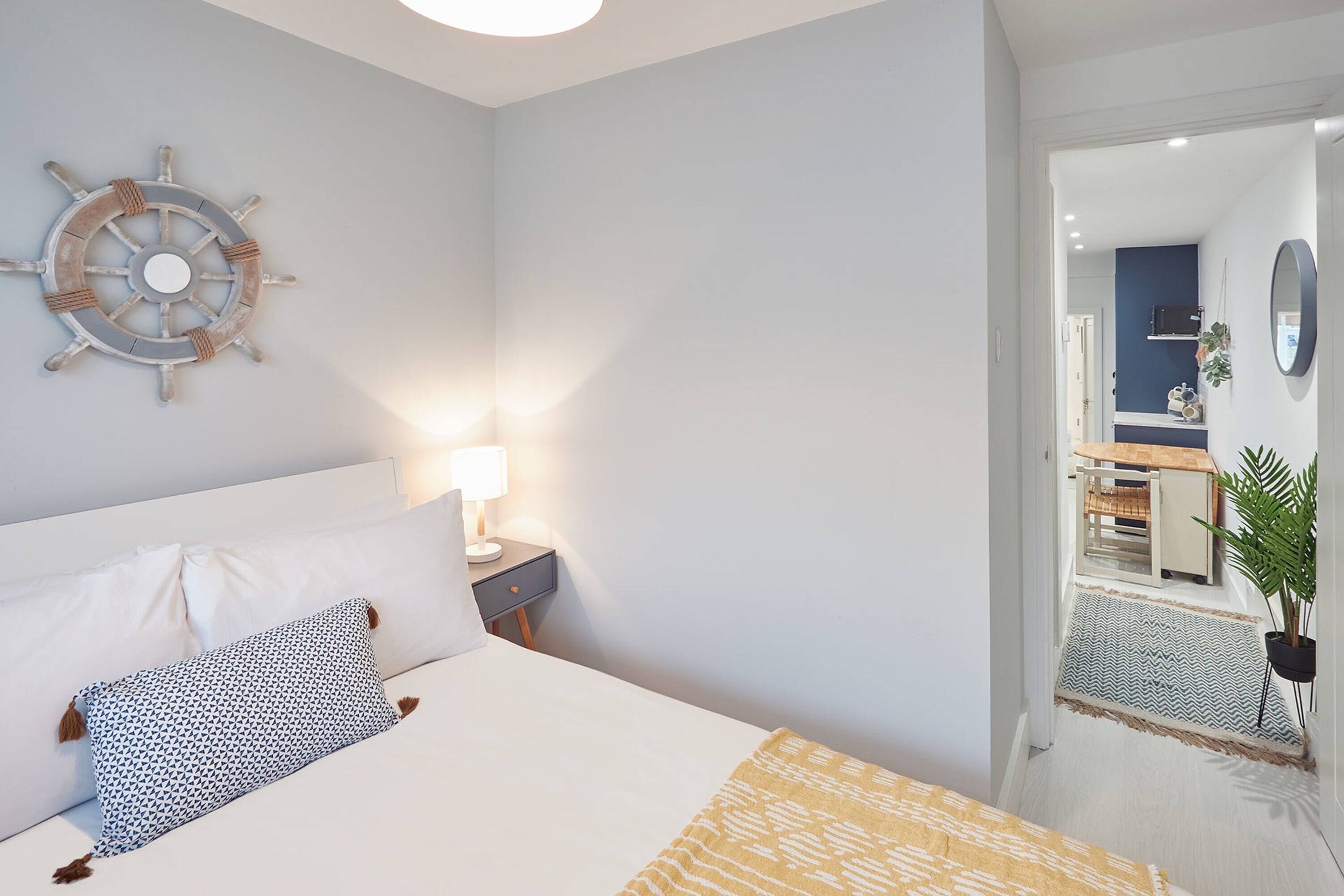 A neatly arranged hotel room with a bed, a wall-mounted ship's wheel, a bedside lamp, and a view into the adjacent room.
