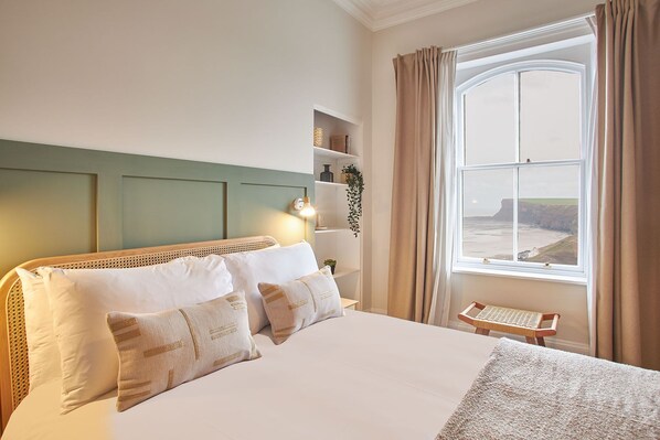 A neatly arranged bedroom with a large bed, a bedside table, a small chair, and a window offering a view of cliffs. - Host Stay Huntcliff View Apartment (Saltburn-by-the-Sea)