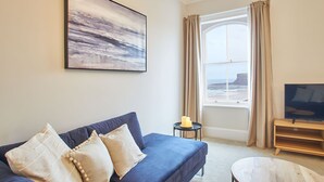 Un salon avec un canapé bleu, des coussins beige, un tableau, une télévision et une fenêtre donnant sur la mer.
