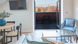 Ett modernt vardagsrum med en soffa, ett soffbord i glas och en matplats med stolar och ett bord. Det finns ett stort fönster med utsikt över byggnader och en balkong med stolar.