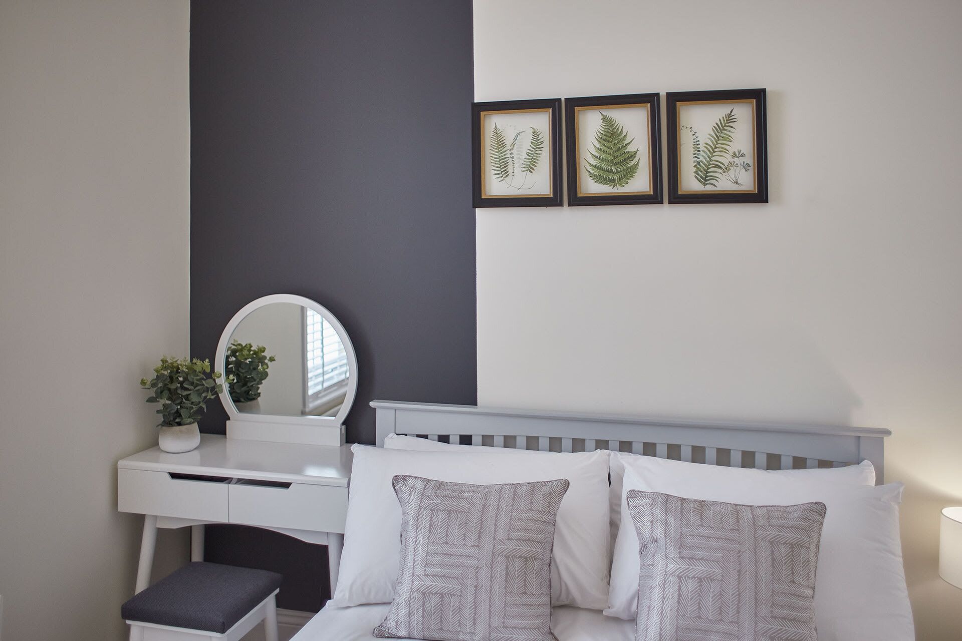 A bedroom with a white bed, two pillows, a bedside table with a mirror, and three framed botanical prints on the wall.