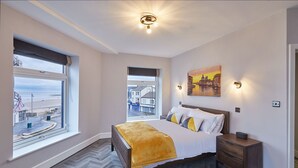 A bedroom with a large bed, a nightstand, a window with a view of a beach and buildings, and a painting of a cityscape.