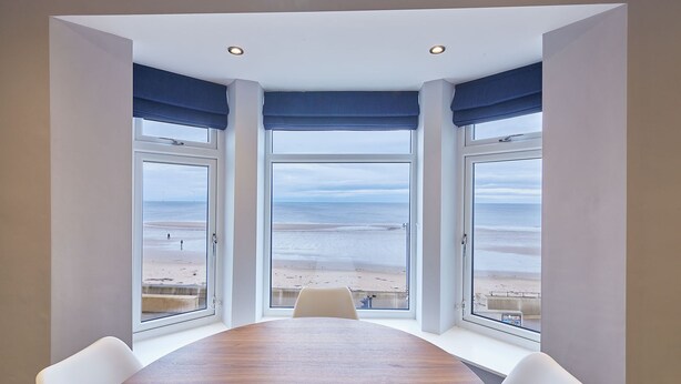 A conference room with a large table and four chairs, large windows with blue curtains, and a view of the beach.