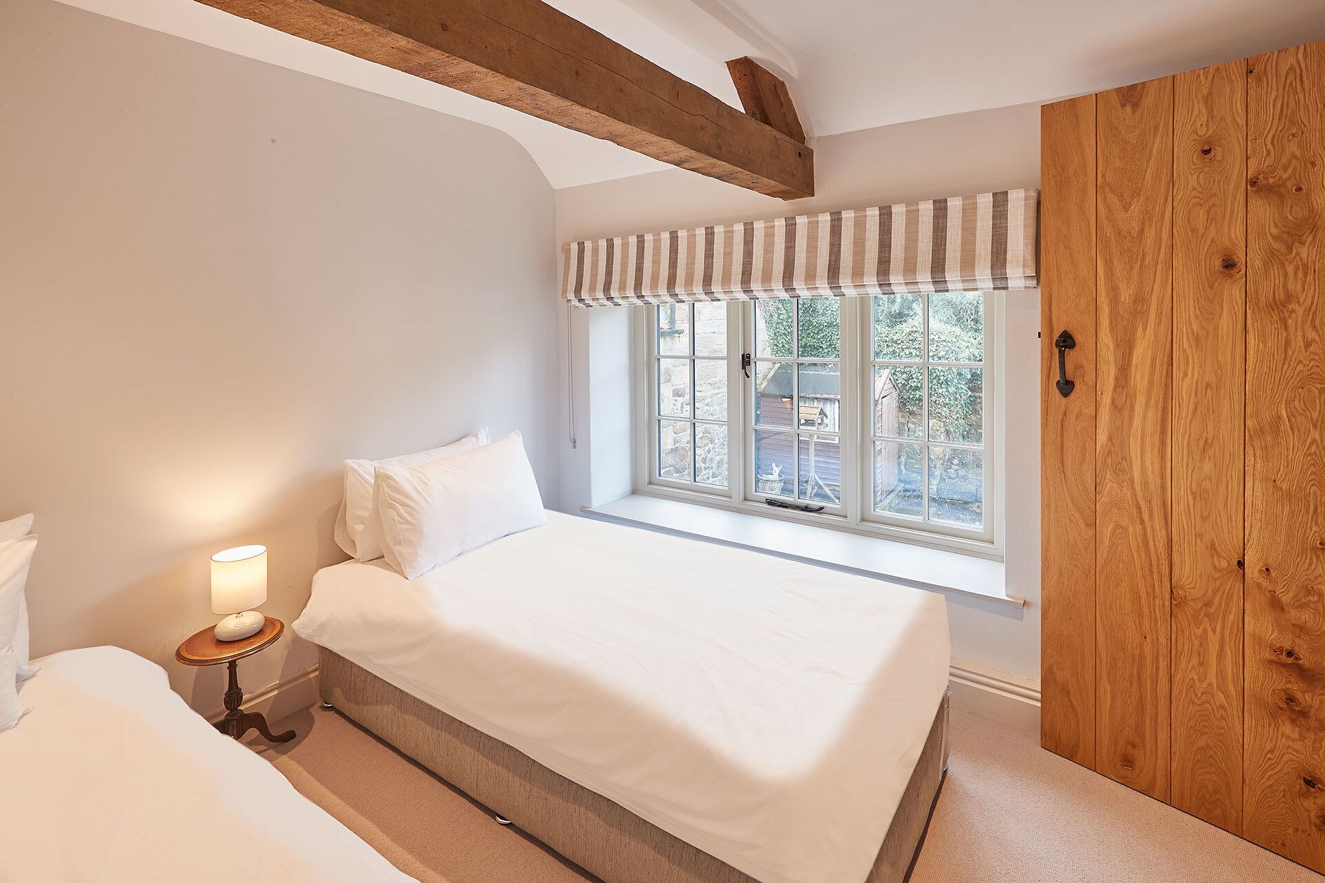 Un dormitorio con una cama grande, una mesita de noche con lámpara, una ventana con vistas al exterior y vigas de madera en el techo.