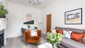 A modern living room with a grey sofa, orange armchair, and a white coffee table. There is a fireplace, a dining area, and a large window with a view of the sea. - Host Stay Feversham House (York)