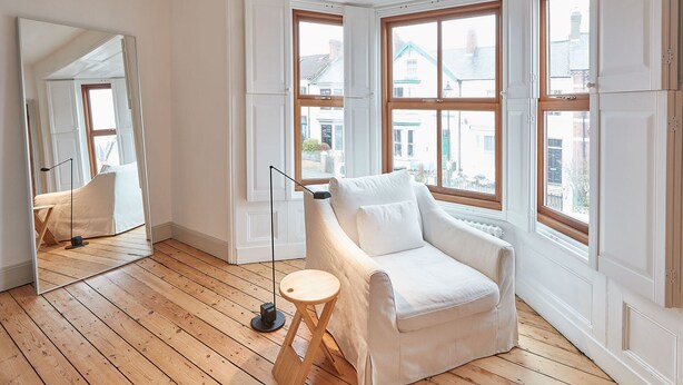 Ein Zimmer mit Holzboden, einem weißen Sessel, einem kleinen Hocker und einem Spiegel, der den Raum widerspiegelt.