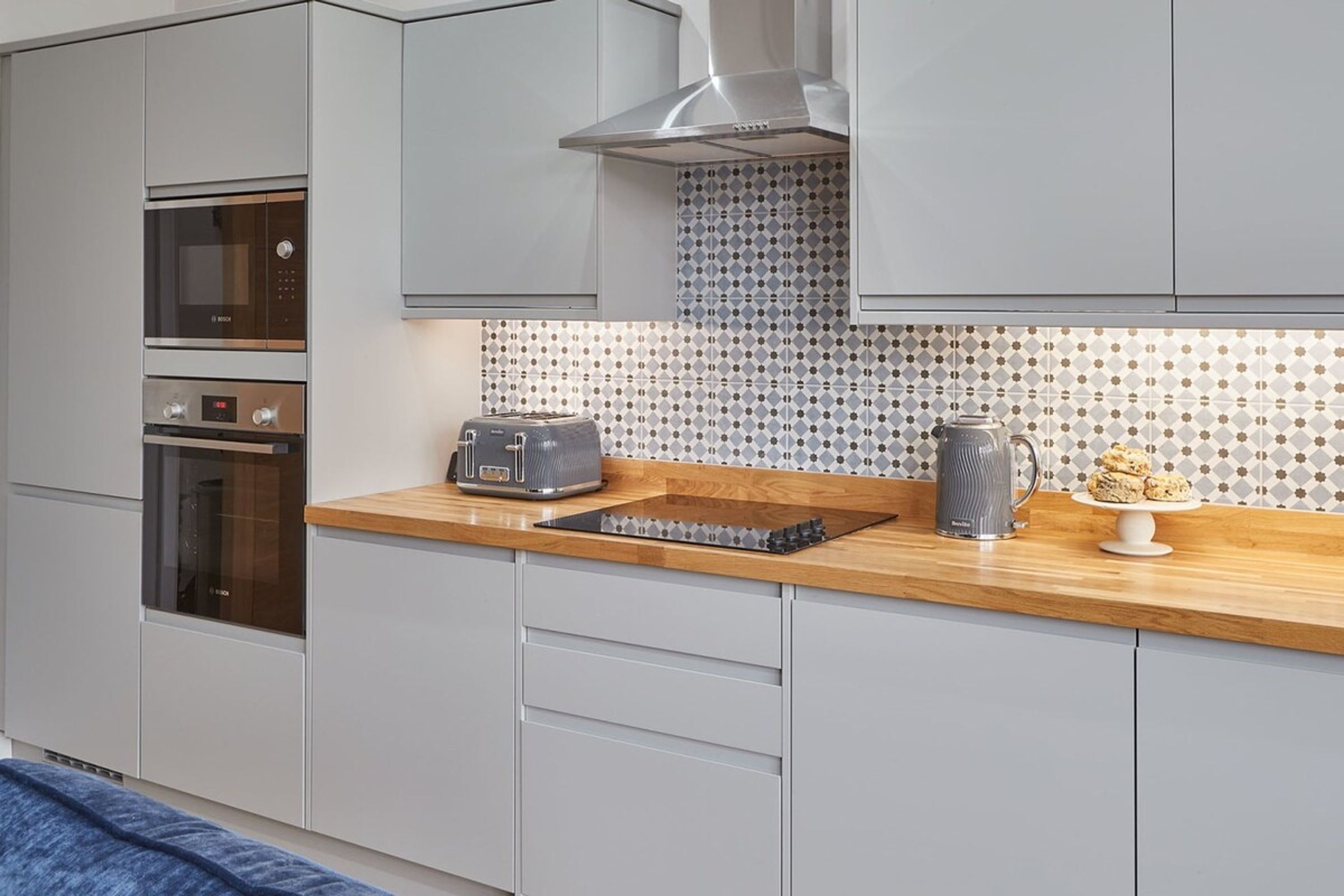 A modern kitchen with grey cabinets, a stainless steel range hood, a microwave, an oven, a toaster, and a coffee maker.