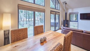 A spacious dining area with a wooden table and woven chairs, large windows offering a view of trees, and a fireplace in the background. - Host Stay Simon Howe Lodge (Whitby)
