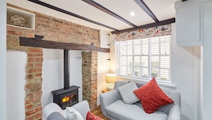 A cozy living room with a brick wall, a fireplace, a sofa with red and grey cushions, and a window with blinds. - Host Stay Thimble Cottage (Whitby)