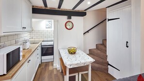 A compact kitchen with white cabinets, a microwave, a toaster, and a small oven. There's a wooden table with a basket of fruit and a clock on the wall. - Host Stay Thimble Cottage (Whitby)