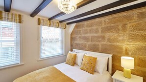 A bedroom with a stone wall, a bed with white bedding and yellow pillows, a bedside table with a lamp, and a window with blinds. - Host Stay Thimble Cottage (Whitby)