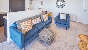 A modern living room with a blue sofa set, a matching armchair, and a patterned ottoman. - Host Stay The Waves (Whitby)