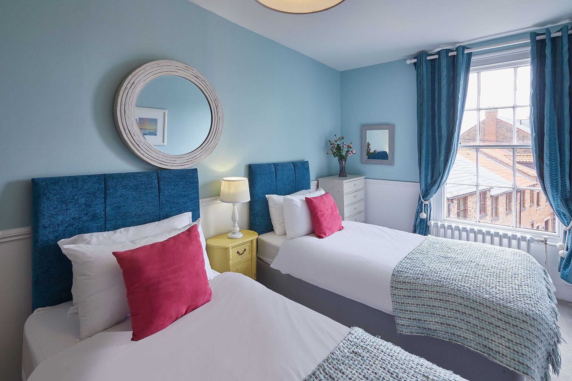 A hotel room with two beds, a round mirror, a bedside table with a lamp, a dresser, and a window with curtains.