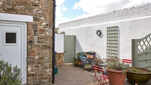 A stone building with a white door, a clock on the wall, and a gravel courtyard with potted plants and a red chair. - Host Stay Wood Street Cottage (Barnard Castle)