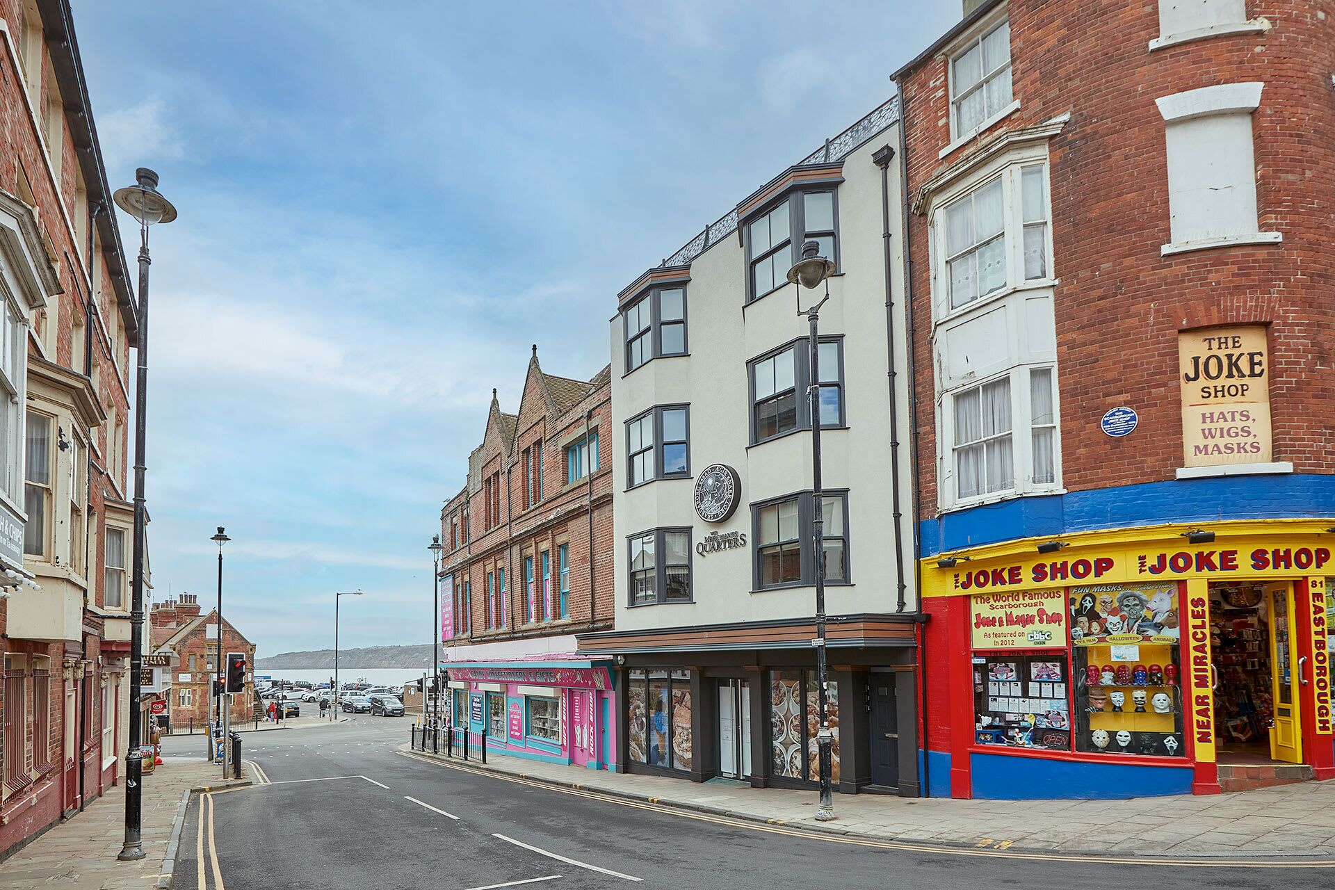 A street view with buildings, including a shop named 'The Joke Shop' selling hats, wigs, and masks.