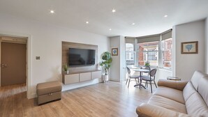 Ein modernes Wohnzimmer mit einem großen Flachbildfernseher, einer Couch, einem Essbereich mit Stühlen und Blick ins Freie.