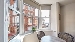 Ein Zimmer mit Esstisch und Stühlen, ein Fenster mit Blick auf ein Backsteingebäude und eine kleine Pflanze auf dem Fensterbrett.