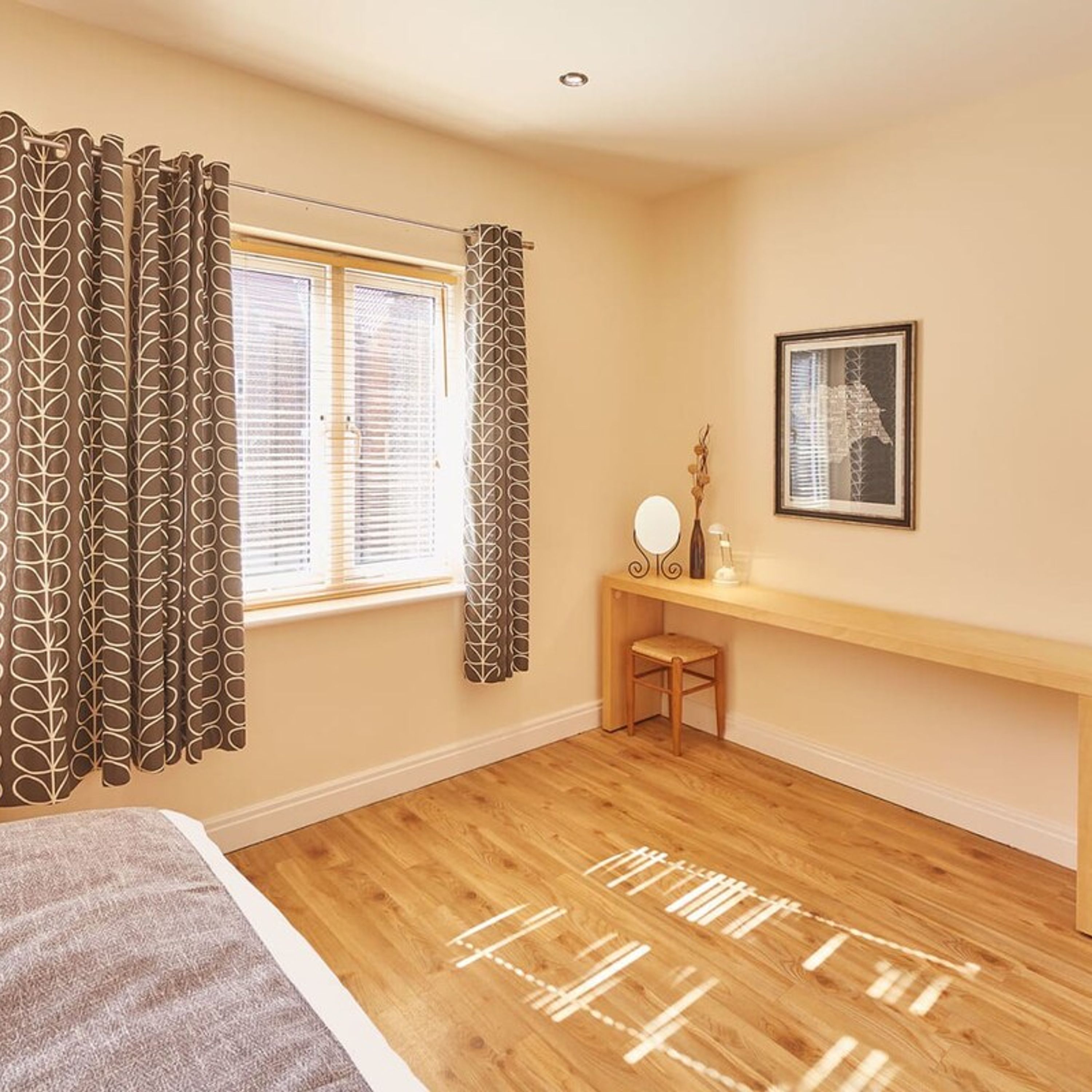 A bedroom with a wooden floor, a bed, a bedside table with a mirror, and a window with blinds.