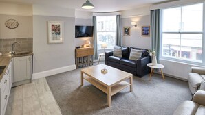 A modern living room with a sofa, coffee table, and a TV. There is a kitchen area with a sink and cabinets. The room has large windows with curtains.
