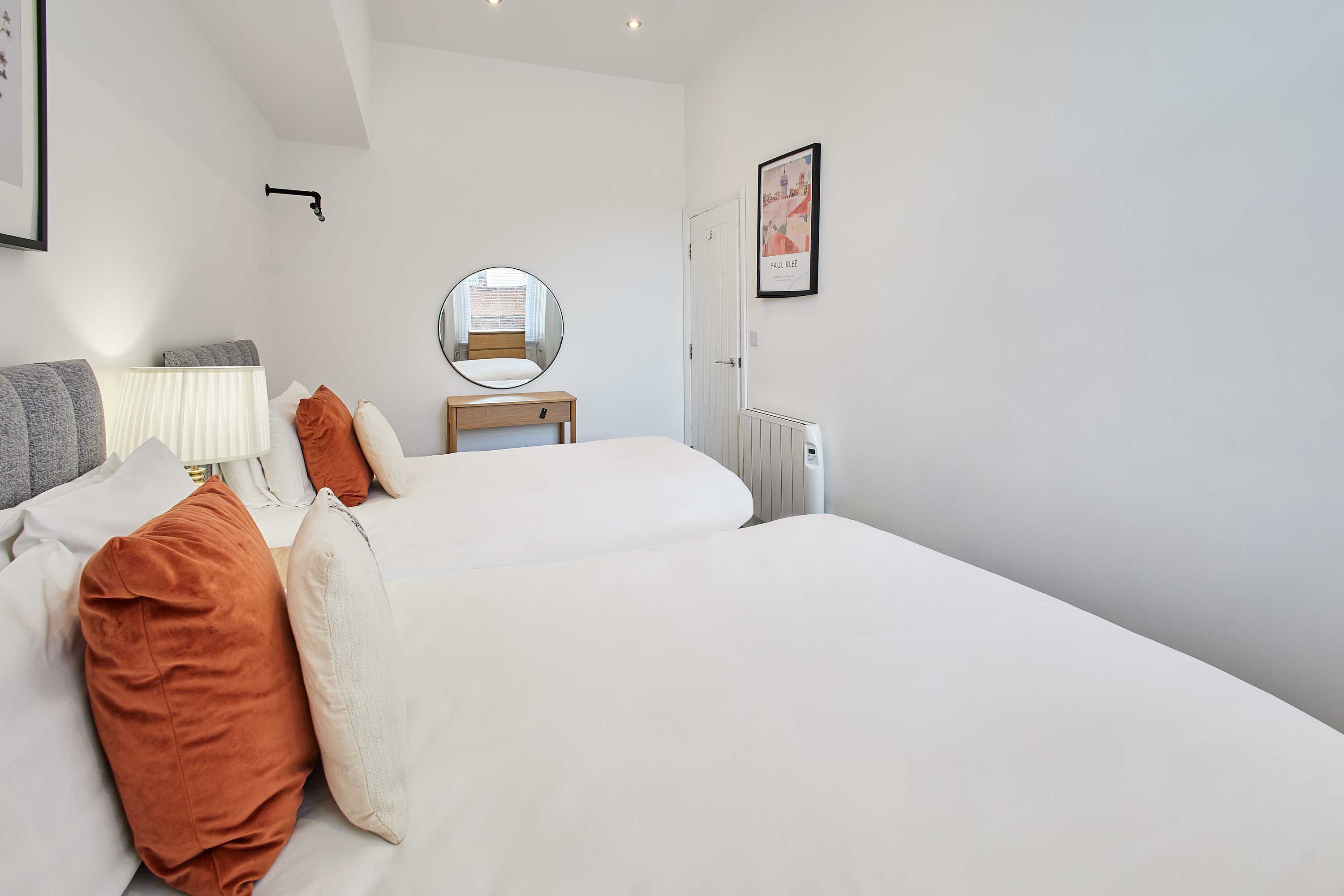 A bedroom with two beds, a bedside table, a lamp, a mirror, and a framed picture on the wall.