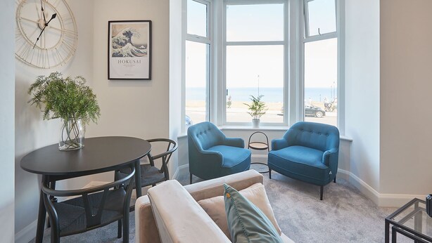 A modern living room with a round table, two blue armchairs, a beige sofa, a large clock, and a framed artwork on the wall. - Host Stay Seaview Retreat (Whitby)
