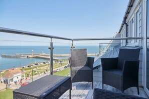 A balcony with wicker furniture overlooking a coastal area, including a lighthouse and a pier.