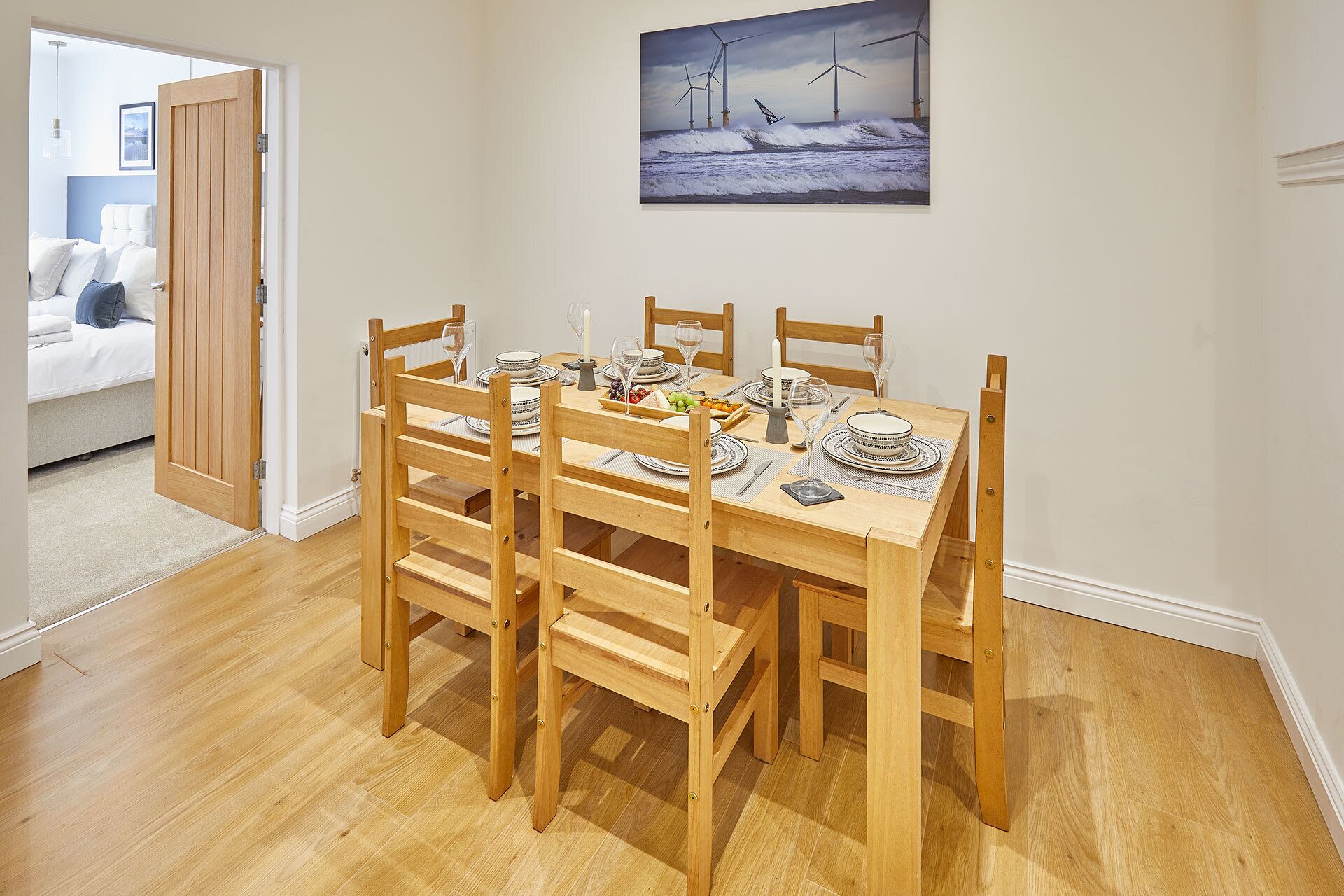 A dining area with a wooden table set for six, surrounded by six wooden chairs. A painting of wind turbines is on the wall above the table.