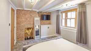 A bedroom with a brick wall, a fireplace, a large bed, a TV mounted above the fireplace, and a window with curtains. - Host Stay 11 Bakehouse Yard (Whitby)