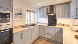 A modern kitchen with grey cabinets, a black range hood, a built-in oven, a sink, and a window with blinds. - Host Stay Sea Watcher (Whitby)